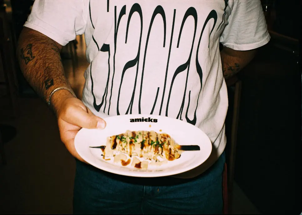 A waiter in a T-shirt that reads Gracias holds a celeriac skewer on a Bar Amicks plate