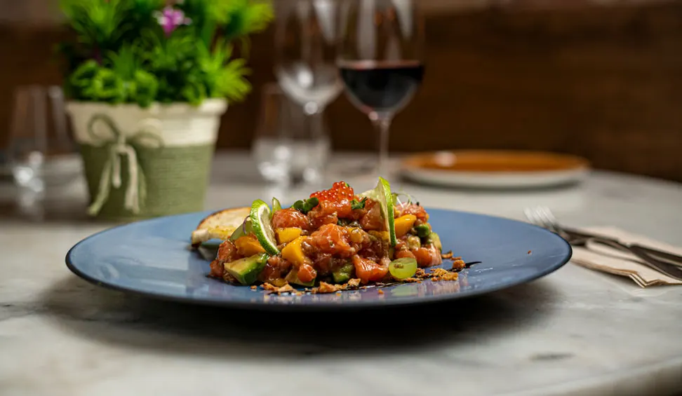 Tàrtar amb mango i alvocat en un plat blau, amb una copa de vi negre al darrere