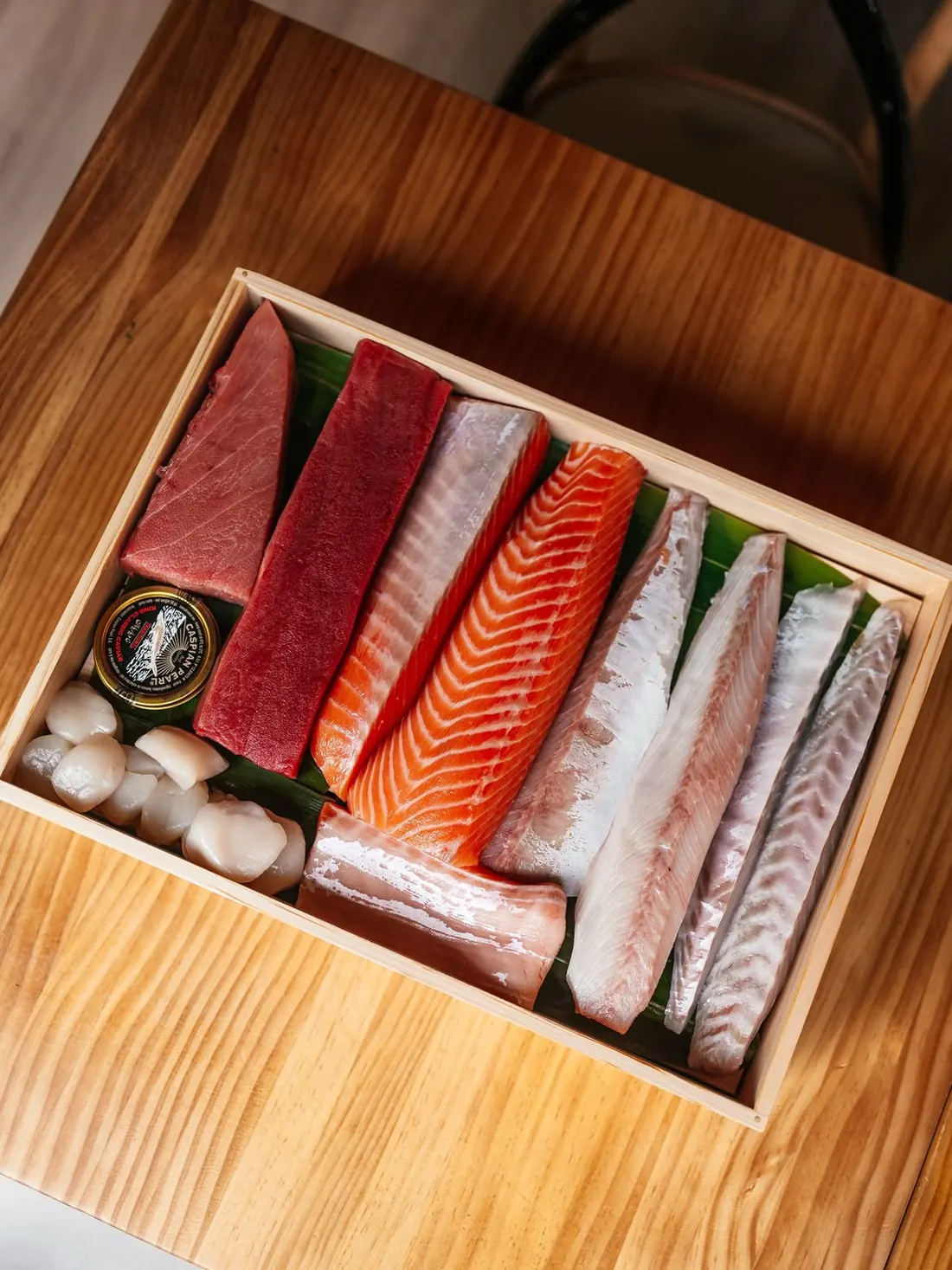 Caja de madera con lomos de atún, salmón y pescado blanco, vieiras y una lata de caviar
