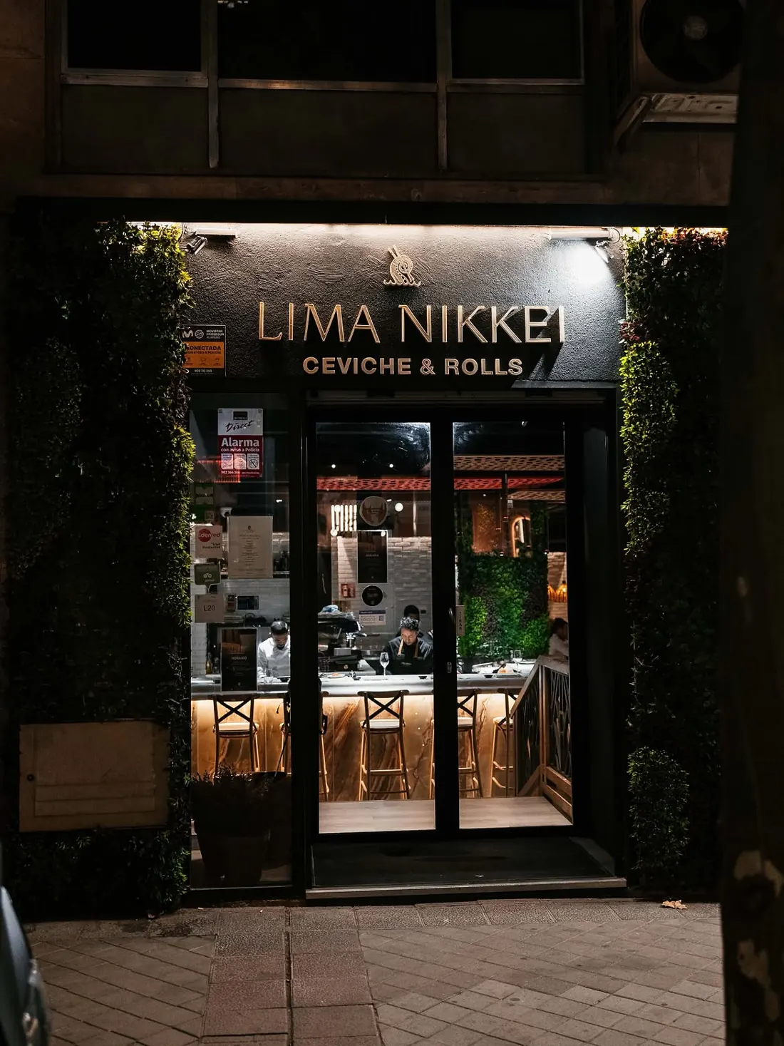 La fachada de Lima Nikkei de noche, con el rótulo «Ceviche & Rolls» y la barra al fondo