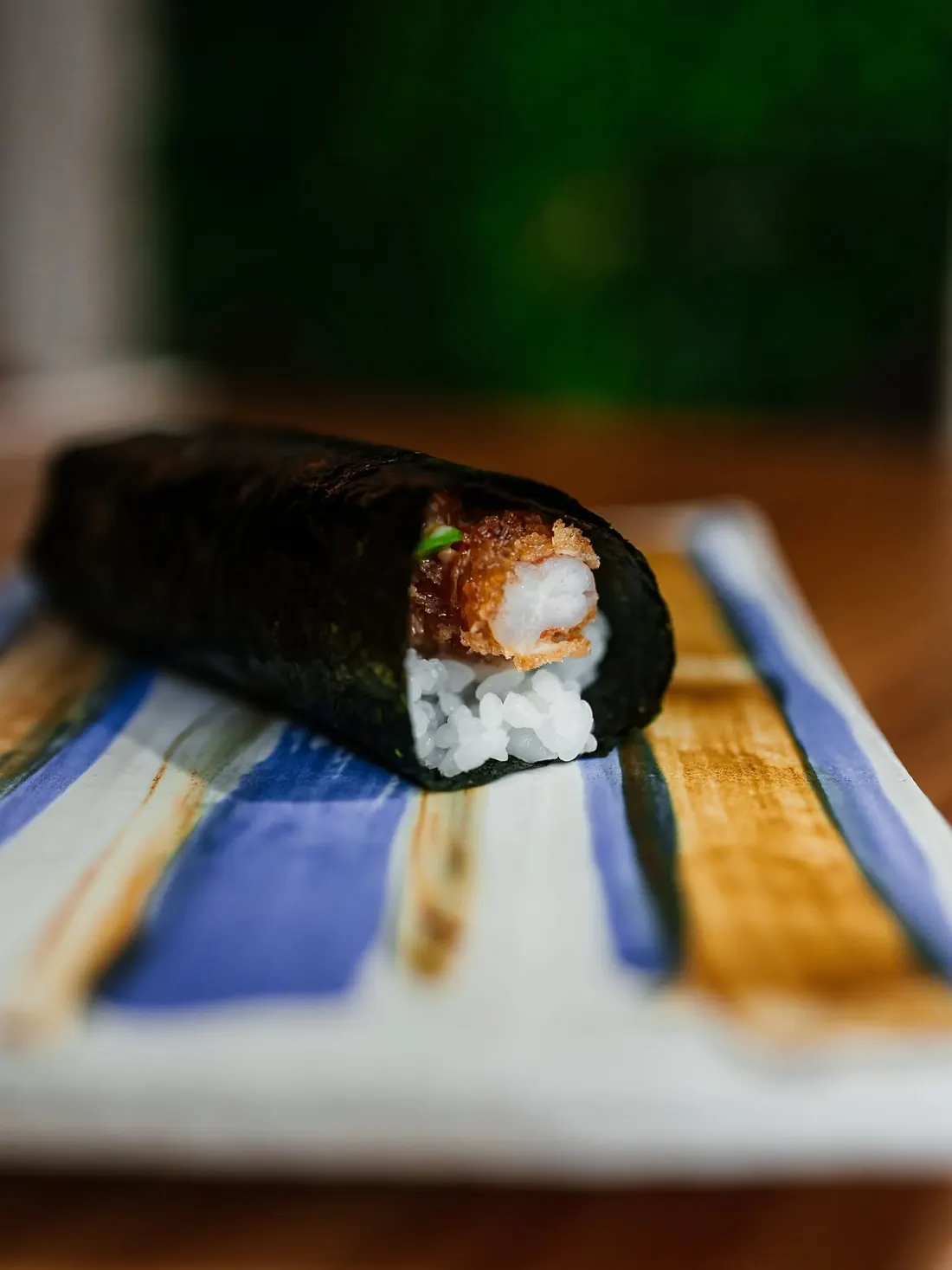 Un hand roll de langostino crujiente envuelto en alga nori, sobre un plato pintado a rayas